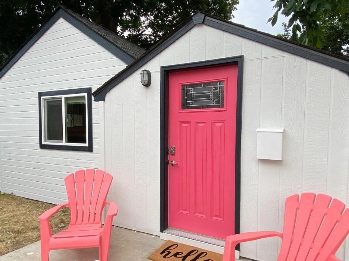 A finished white cottage with a bold coral door and matching chairs on the porch